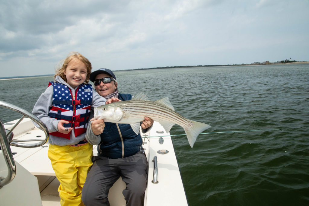young girl in red white and blue life jacket pats fish held by Fly Girl and guide Capt Avery Revere