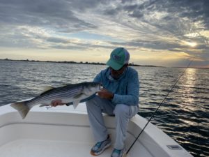 man in a blue shirt and blue hat holding a striped bass with swirling clouds in the backgroudn