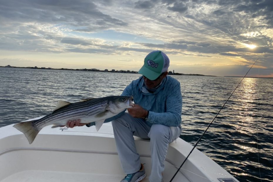 man in a blue shirt and blue hat holding a striped bass with swirling clouds in the backgroudn
