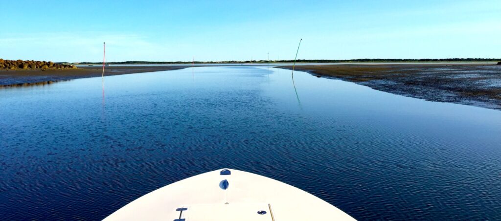 Bow of boat leaving Barnstable Harbor marina channel with mudflats on either side of channel.