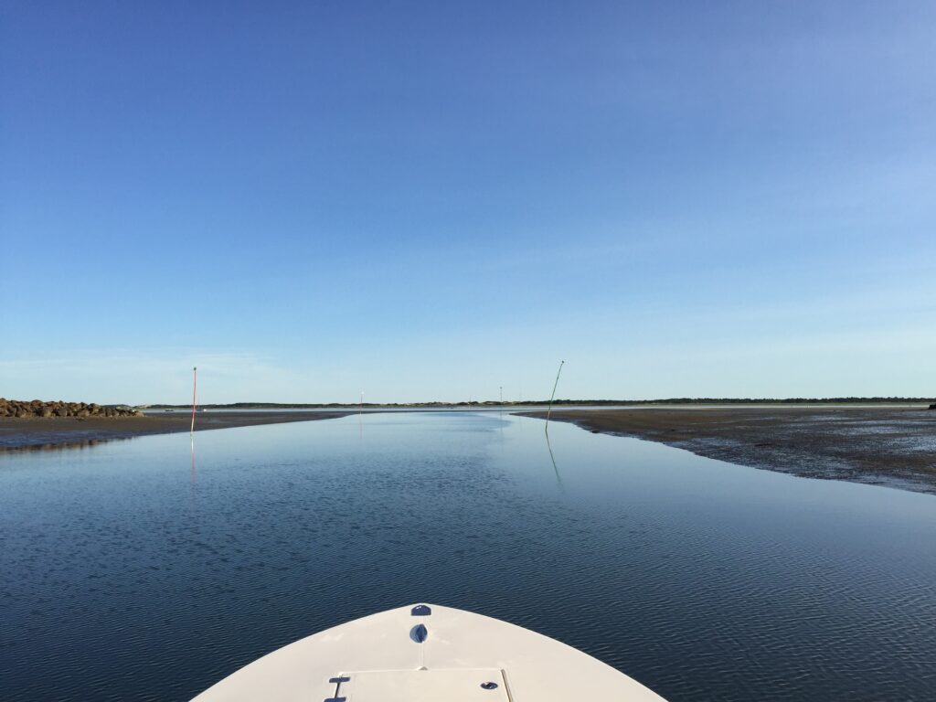 View over the bow of a boat of a channel with sandflats on either side and channel markers